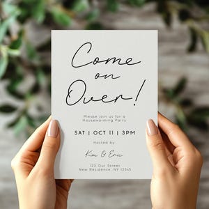 May include: A housewarming party invitation held by hands. The invitation is a light gray rectangle with black script that reads "Come on Over!" and details for the event. The background is blurred with green foliage.