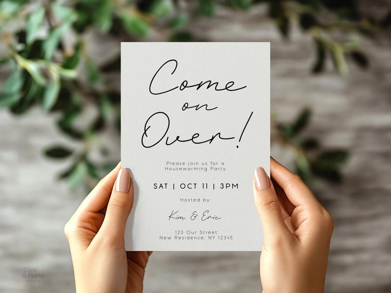 May include: A housewarming party invitation held by hands. The invitation is a light gray rectangle with black script that reads "Come on Over!" and details for the event. The background is blurred with green foliage.