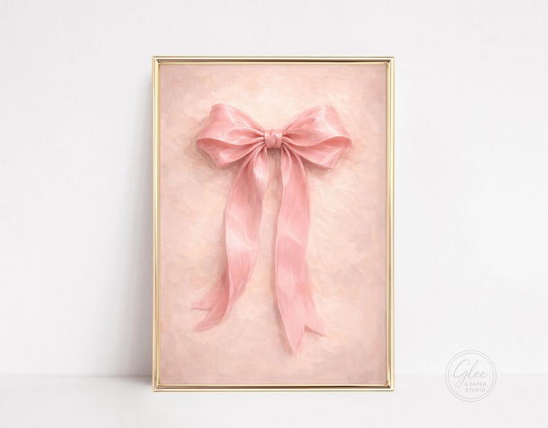 May include: A framed print featuring a large, pink bow with long, flowing ribbons. The bow is the focal point, set against a soft, textured background. The frame is gold. The print is from Glee Paper Studio.