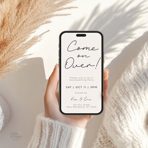May include: A smartphone displays a housewarming party invitation with the text "Come on Over!" in elegant script. The invitation includes the date, time, and host's names. The phone is held against a soft, neutral background with dried plants.