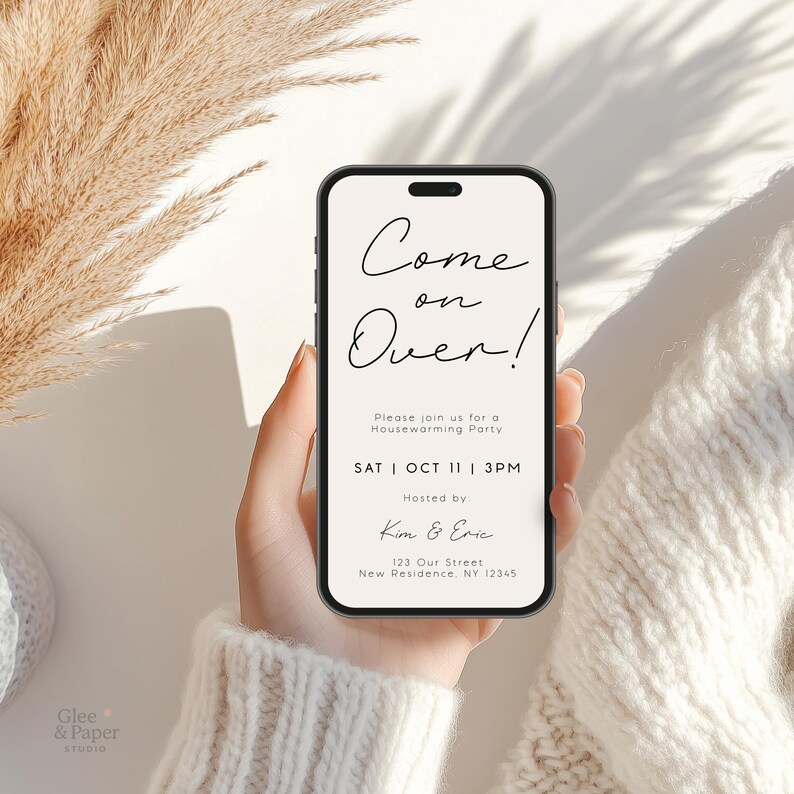 May include: A smartphone displays a housewarming party invitation with the text "Come on Over!" in elegant script. The invitation includes the date, time, and host's names. The phone is held against a soft, neutral background with dried plants.