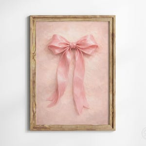 May include: A framed print featuring a large, soft pink bow with long, flowing ribbons. The bow is centered on a light pink, textured background. The wooden frame adds a rustic touch to the artwork.