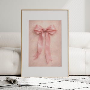 May include: A framed print featuring a soft pink bow with long, flowing ribbons. The bow is centered on a textured, light pink background. The frame is gold, and the print is displayed on a white couch and a patterned rug.