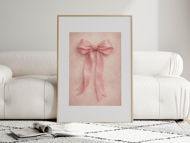 May include: A framed print featuring a soft pink bow with long, flowing ribbons. The bow is centered on a textured, light pink background. The frame is gold, and the print is displayed on a white couch and a patterned rug.