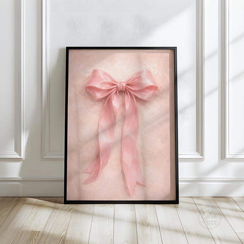 May include: A framed art print featuring a large, soft pink bow with long, flowing ribbons. The bow is centered on a light pink background, and the artwork is encased in a black frame. The print is displayed against a white wall.