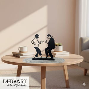 May include: A black metal sculpture depicting two people dancing, placed on a wooden table. The figures are stylized silhouettes, one in a suit, the other in a blouse and trousers. The sculpture sits on a gray surface. The text "DERVART - dekoratif ürünler" is at the bottom.