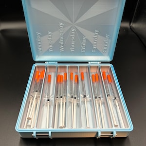 May include: A light blue weekly pill organizer with a hinged lid, labeled with days of the week. Inside are multiple syringes with orange caps. The organizer is open, revealing the contents.