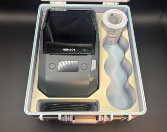 May include: A dark gray Niimbot label printer with a black screen and control buttons, housed in a light-colored carrying case. The case includes a roll of white label tape, a black charging cable, and custom-molded compartments for storage.