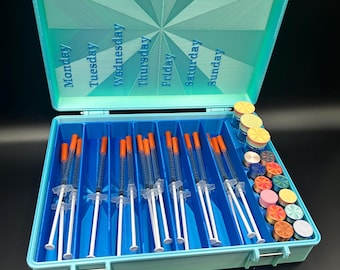 May include: A light blue plastic storage case with a hinged lid featuring a sunburst pattern and the days of the week printed in blue. The interior contains seven vertical compartments holding clear syringes with orange caps and small vials.