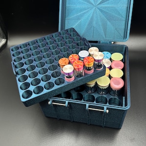 May include: A teal storage box with a geometric lid, open to reveal small glass vials with colorful lids. The box has a tray with multiple circular openings to hold the vials. The box is on a dark surface.