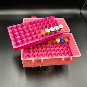 May include: A pink plastic storage case with a removable tray, designed to hold small vials. The case is open, revealing multiple vials of various colors. The case has a handle and latches for secure closure.