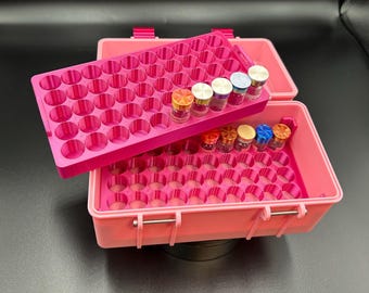 May include: A bright pink plastic storage case featuring a removable tray with multiple circular slots. The tray and the base of the case are filled with small, clear glass vials topped with metallic caps in various colors including orange, blue, and silver.