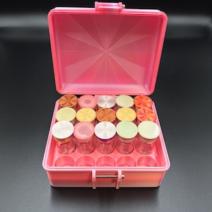 May include: A pink plastic container with a hinged lid, open to reveal small glass jars with various colored lids. The container is on a black surface. The lids are in shades of gold, pink, and green.