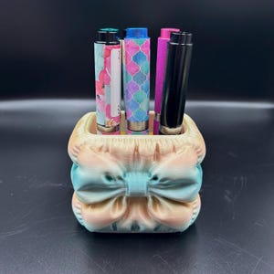 May include: A decorative pen holder with a bow design in pastel colors, holding several pens with colorful patterns. The pens include floral, geometric, and solid colors. The holder is approximately 3 inches wide and 3 inches tall.