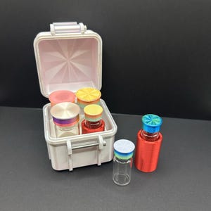 May include: A white miniature case with a pink interior, holding several small glass vials with colorful lids. Two vials are outside the case, one with a red holder and a blue and green lid, and another with a white and blue lid.
