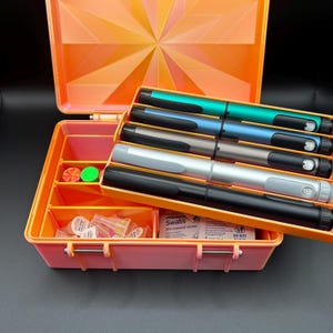 May include: An open orange plastic storage case with a geometric lid pattern. Inside are five pens in teal, blue, grey, silver, and black, plus small containers and alcohol swab packets. The case features dividers and a hinged design.