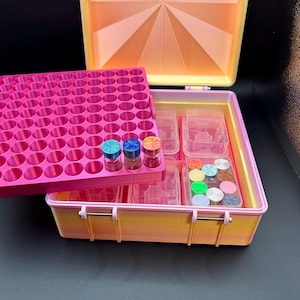 May include: A rectangular storage case with a gradient yellow and pink exterior features a removable bright pink tray with numerous circular slots. Three small glass vials with textured metallic caps in blue, teal, and orange sit on the tray.