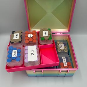May include: A multi-tiered, rainbow-colored storage box with a hinged lid, containing several clear plastic containers filled with small, colorful circular items. Each container features a white label with text including SK10, 50AM, 01XS, TR40, RT30, R30, and 10AM YOYO.