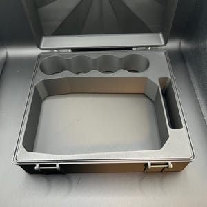 May include: A black rectangular storage case with a hinged lid featuring a sunburst pattern. The interior contains custom-molded foam inserts with four circular cutouts along the top edge, a large central rectangular compartment, and a side slot.