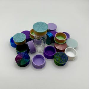 May include: A collection of colorful, small, round plugs in various shades of blue, green, purple, and pink. A clear glass vial is in the center of the plugs. The plugs have a variety of textures and patterns.