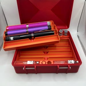 May include: A red plastic case with an orange interior, holding several medical devices. The case contains three pens in pink glitter, purple, and black, along with small glass vials. The case is open, revealing multiple compartments.