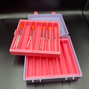 May include: A pink plastic organizer with a lavender lid, containing several syringes with orange caps. The organizer is open, revealing the syringes neatly arranged in individual slots. The text "AMI" is visible on the organizer.
