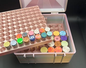 May include: A light pink storage case with a multi-tiered grid insert for organizing small cylindrical containers. The case features a hinged lid and contains numerous small jars with colorful, patterned, and metallic circular lids.