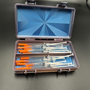 May include: A rectangular, light purple storage case with a hinged lid and a blue interior featuring a sunburst pattern. The case contains multiple clear plastic syringes with orange caps and white plungers, labeled with the text Easy-Touch.