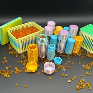May include: A collection of small, cylindrical, perforated containers in various colors including yellow, blue, and lavender, alongside two rectangular, slatted storage boxes. The containers are filled with small, translucent orange beads.