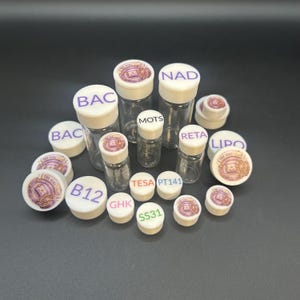 Könnte beinhalten: Eine Sammlung von klaren Glasfläschchen mit weißen Verschlüssen, einige mit aufgedruckten Etiketten wie BAC, NAD, MOTS, RETA, LIPO, B12, GHK, TESA, PT141 und SS31. Mehrere Verschlüsse zeigen ein kreisförmiges Logo mit dem Text The Vial Vault.