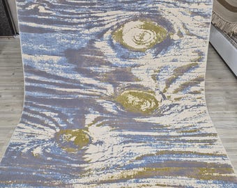 May include: A rectangular area rug with a wood grain design. The rug features a pattern of blue, gray, and beige hues, with circular knot-like details in shades of green and yellow. The rug has a fringed edge.