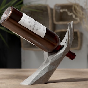 Könnte beinhalten: Eine braune Weinflasche mit weißem Etikett wird in einem grauen, geometrischen, handförmigen Weinflaschenhalter gehalten. Der Halter besteht aus Beton und steht auf einer Holzoberfläche. Die Flasche ist angewinkelt und zeigt das Etikett.