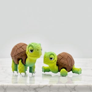 Könnte beinhalten: Zwei bewegliche Schildkrötenfiguren mit hellgrünen Köpfen und Gliedmaßen sowie braunen, strukturierten Panzern. Die Schildkröten haben große weiße Augen mit schwarzen Pupillen und stehen nebeneinander, eine aufrecht und die andere kriechend.