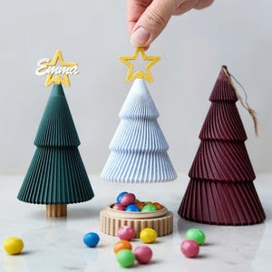 Könnte beinhalten: Drei kegelförmige Weihnachtsbaum-Behälter mit plissierter Textur in Dunkelgrün, Hellblau und Dunkelrot. Der grüne Baum hat einen goldenen Sternaufsatz mit dem Namen Emma. Der blaue Baum wird angehoben und gibt eine kleine runde Basis voller bunter Bonbons frei.