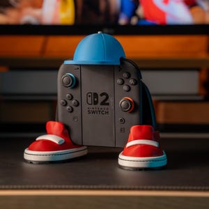 Könnte beinhalten: Ein Nintendo Switch-Controller mit einer blauen Baseballkappe und roten Turnschuhen. Der Controller ist schwarz mit weißen Knöpfen und dem Text "Nintendo Switch". Die Turnschuhe haben weiße Sohlen und Schnürsenkel.