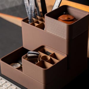 Könnte beinhalten: Ein brauner Schreibtisch-Organizer mit mehreren Fächern und Ebenen. Der Organizer enthält Stifte, kleine Accessoires und einen kleinen Lederbeutel. Das Design ist modern und funktional, geeignet für Büro oder Zuhause.