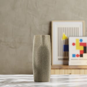Könnte beinhalten: Eine hohe, strukturierte Vase in einem neutralen Beigeton. Die Vase hat eine zylindrische Form mit leicht breiterem Boden und schmaler Öffnung. Dekorative Kunstdrucke befinden sich im Hintergrund.