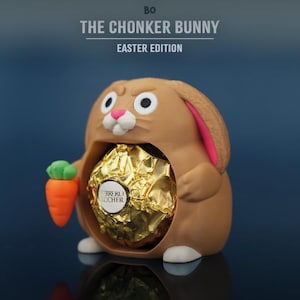 Könnte beinhalten: Eine braune "Chonker Bunny" Oster-Edition-Figur, die eine goldfarben verpackte Schokoladenbonbons hält. Der Hase hat rosa Innenohren, weiße Akzente und eine kleine orangefarbene Karotte. Der Text oben lautet "Bo" und "THE CHONKER BUNNY EASTER EDITION."