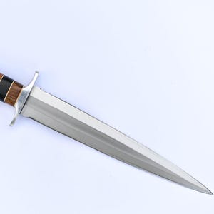 May include: A dagger with a polished steel blade and a handle featuring alternating bands of dark wood and black material. The handle has a silver-colored metal bolster and pommel. The blade is double-edged and tapers to a sharp point.