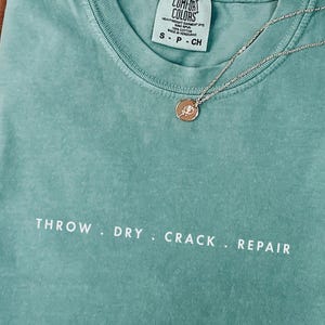 May include: A light green short-sleeve t-shirt with a crew neckline and the text THROW . DRY . CRACK . REPAIR printed in white across the chest. The shirt features a Comfort Colors brand label at the inner neck and a thin gold-tone chain necklace.