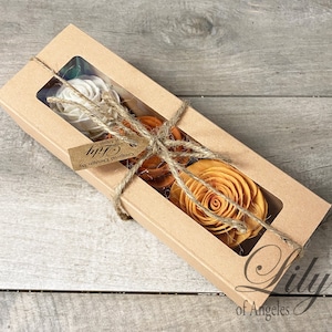 May include: A rectangular kraft paper box with a clear window, tied with twine, containing several wooden flowers in shades of white and orange. A small tag reads "Lily of Angeles".