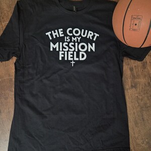 Puede incluir: Camiseta negra con el texto blanco "THE COURT IS MY MISSION FIELD" y una pequeña cruz. Un balón de baloncesto marrón descansa a la derecha de la camiseta, sobre una superficie de madera.