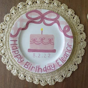 May include: A decorative plate with a pink birthday cake design and the text "Happy Birthday Eleanor 8.2.22". The plate is surrounded by a pink bow design and sits on a woven placemat.