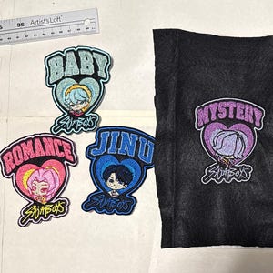May include: Embroidered patches and fabric pieces featuring cartoon characters and text. Patches include "BABY," "ROMANCE," and "JINU" in heart-shaped designs. Two black fabric pieces display "MYSTERY" and "ABBY" patches. A ruler is in the background.