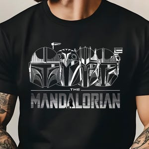 May include: Black t-shirt featuring a graphic of four Mandalorian helmets in white. The text "THE MANDALORIAN" is printed in silver below the helmets. The design is centered on the shirt.