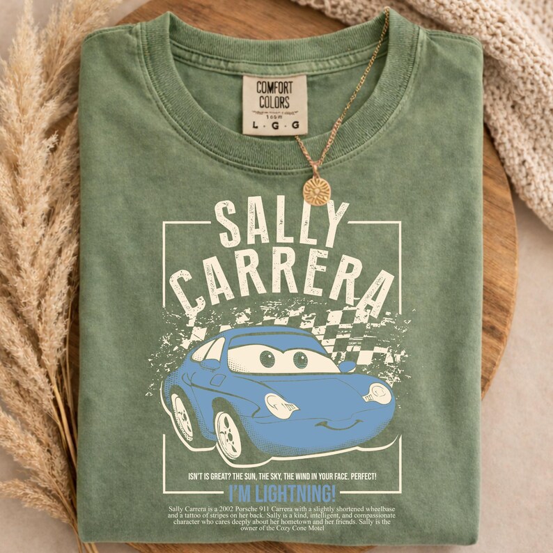 May include: Green Comfort Colors t-shirt featuring a blue cartoon car with the text "SALLY CARRERA" and "I'M LIGHTNING!". The shirt also includes a description of the car and its owner. A gold necklace with a circular pendant is draped over the shirt.