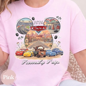 Puede incluir: Camiseta rosa claro con un dise&ntilde;o de la pel&iacute;cula Cars. El dise&ntilde;o incluye el texto "2026 Cars Family Trip" e ilustraciones de pistas de carreras, coches y un paisaje des&eacute;rtico. La camiseta es de color rosa claro.