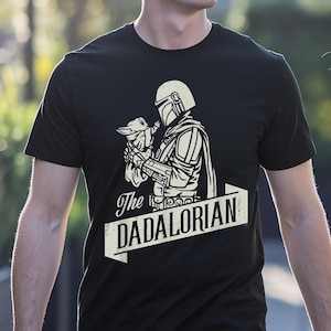 May include: Black t-shirt with a white graphic of The Mandalorian holding Grogu, and the text "The DADALORIAN" in a banner. The design is a stylized illustration with a vintage aesthetic.