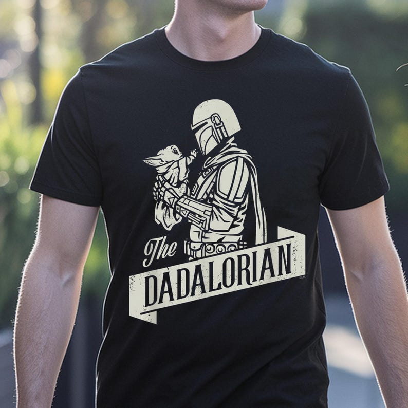 May include: Black t-shirt with a white graphic of The Mandalorian holding Grogu, and the text "The DADALORIAN" in a banner. The design is a stylized illustration with a vintage aesthetic.