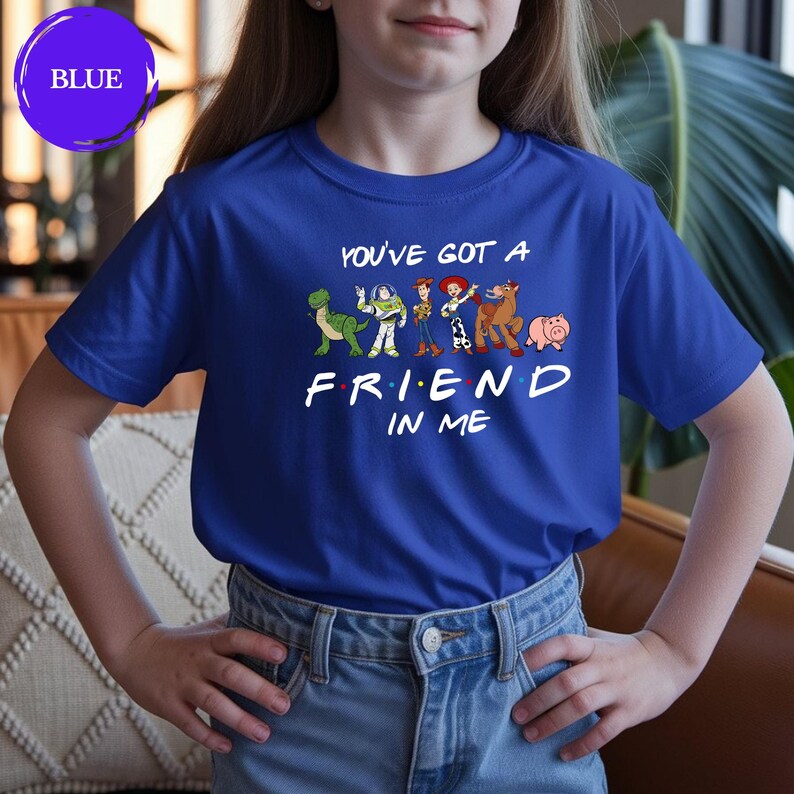 May include: A royal blue t-shirt featuring the phrase "YOU'VE GOT A FRIEND IN ME" with cartoon character graphics. The shirt has short sleeves and is worn by a person. The word "BLUE" is in a circle in the upper left corner.